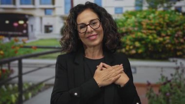 Orta yaşlı, gözlüklü, siyah giysili, elleri dışarıda bina zeminine karşı olan İspanyol bir kadın..