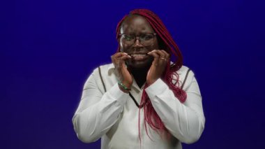 Kırmızı örgülü ve gözlüklü bir kadın mavi arka planda endişeli görünüyor. Endişelerini ya da endişelerini dile getiriyor..