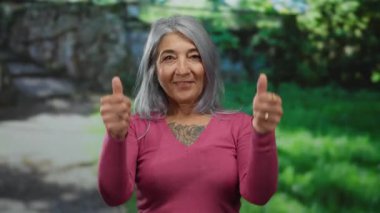 Pembe kazaklı, gri saçlı yaşlı bir kadın. Güneşli bir parkta gülümseyip el sallıyor..