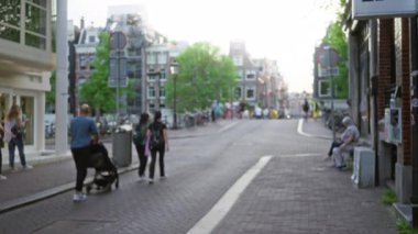 Amsterdam 'da şehir caddesinde yürüyen ve bisiklet süren bulanık insanların olduğu sokak sahnesi, tarihi mimari ve yeşilliğin dağılmış arka planıyla şehir yaşamını gözler önüne seriyor..