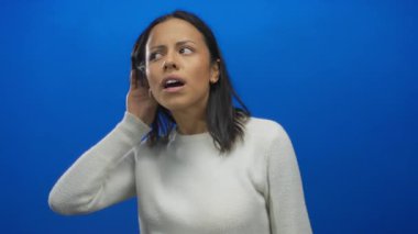 Bir kadın, canlı mavi bir arka plana karşı dikkatle dinliyor. Bir eli kulağında, bir yüzü de etkileyici bir şekilde merakını ve dikkatini veriyor..