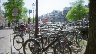 Ağaçların arasından süzülen güneş ışığıyla gölgeli kanallar boyunca park etmiş bisikletler ve arka planda bulanık insanlar mükemmel bir Hollanda manzarası yaratıyor..