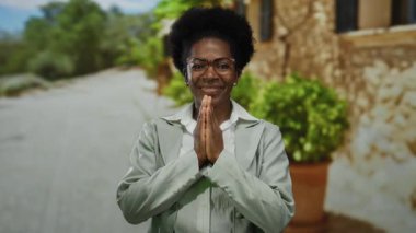 Gözlüklü gülümseyen bir kadın, dua ederken el ele tutuşan, güneşli bir sokakta ayakta duran, arka planda yeşil bir ceket giyen,.