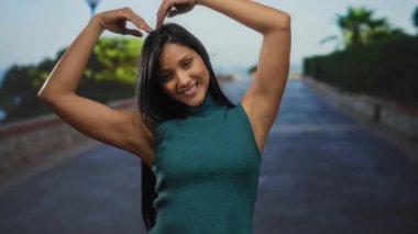 Genç bir kadın ellerini açık havada güneşli bir caddede kalp hareketi yapıyor, gülümsüyor ve canlı bir şehir ortamında mutluluk ve pozitiflik yayıyor..
