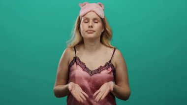 Pembe gecelikli bir kadın, uyku maskesi takmış, sakin bir ortamda yeşil bir zemine yaslanıyor ve nefes alıyor. Rahatlama ve farkındalık sergiliyor..