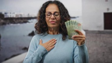 Orta yaşlı, kıvırcık saçlı bir kadın deniz kenarında Arjantin pezosu tutuyor. Arka planda da bir tekne var. Hislerini limanın yanında el üstünde ifade ediyor..