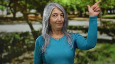 Gri saçlı, mavi kazak giyen son sınıf öğrencisi kadın, açık havada, açık yeşil bir parkta doğal ışık altında sakin bir günün tadını çıkarırken baş parmak işareti yapıyor..