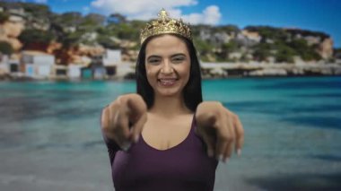 Güneşli plajda, temiz deniz manzaralı arka planda, neşe ve kutlamaların somutlaştırıldığı, tacından hareketler yapan gülümseyen bir kadın..