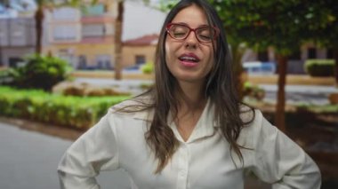 Kırmızı gözlüklü bir kadın binanın önünde elleri kalçasında dilini dışarı çıkartıyor; oyunculuk, eğlence, neşe, spontanelik..