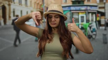 Açık havada şapka giyen genç bir kadın şehir arkaplanına karşı neşeli bir ifadeyle baş parmak kaldırıyor ve baş parmak işareti yapıyor..