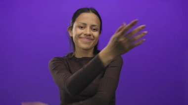 Hispanik kadın, kendini izole edilmiş mor bir arka plana karşı neşeli bir şekilde kucaklıyor, modern bir ortamda özgüvenini ve özgüvenini ifade ediyor..