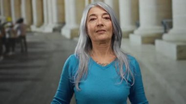 Gri saçlı, mavi kazaklı yaşlı bir kadın arka planda sütunları olan açık bir sokakta kendini işaret ediyor..