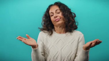 Kıvırcık saçlı, süveter giyen olgun bir kadın canlı mavi bir arka plana karşı güvenli bir şekilde poz verir.