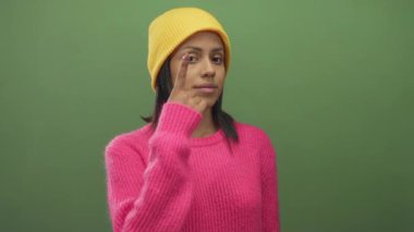 Sarı bereli ve pembe kazaklı genç bir kadın yeşil bir arka plana karşı gözlerini dikmiş canlı ve ilgi çekici bir görsel karşıtlık yaratıyor..