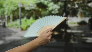 Parkta mavi yelpaze tutan adam güneşli bir günde yeşil arka planı bulanık geleneksel aksesuar sergiliyor..