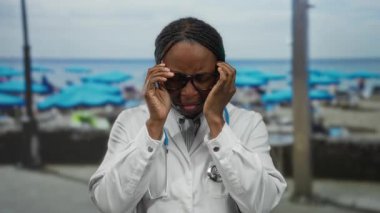 Gözlüklü kadın doktor plajda steteskoplu beyaz bir ceket giyerek güneşli gökyüzünün altında stresli duruyor..
