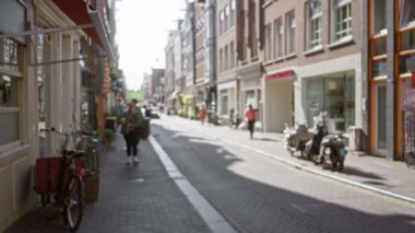 Bulanık Amsterdam caddesinde bisiklet ve scooterlarla gezen bir kadın canlı şehir ortamında güneşli kırsal yolda ilerliyor..