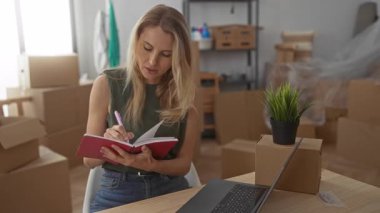 Oturma odasındaki not defterine yeni ev ortamını ve planlama sürecini gösteren kutular ve dizüstü bilgisayarlarla çevrili bir kadın yazıyor..