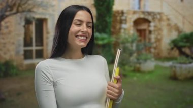 Dışarıdan gülümseyen bir kadın, taştan duvarları ve yemyeşil bahçeleri olan taşralı bir binanın yakınındaki kitapları göstererek ve tutarak neşeli bir akademik sahne yakalıyor..