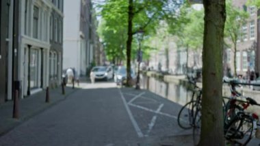Güneşli bir günde bisiklet, araba ve yayalarla Amsterdam Kanal Caddesi 'nin bulanık görüntüsü; ikonik Hollanda mimarisi gösteren kentsel manzara.