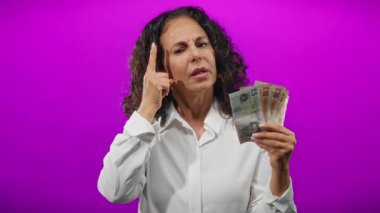 İngiliz banknotlarını ihtiyatlı bir şekilde mor arka planda tutan kadın, izole edilmiş renkli duvarlar üzerinde mali kararları düşünen orta yaşlı bir kadını gösteriyor..