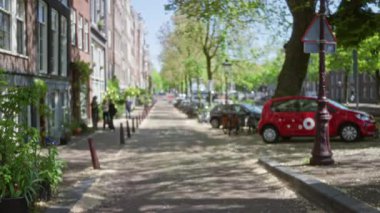 Amsterdam 'da bulanık bir şehir manzarası var. Klasik tuğla binaların ve park halindeki kırmızı bir arabanın yanında yürüyen insanlarla dolu..