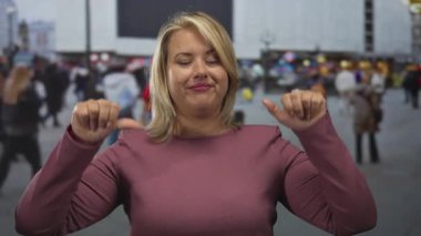 Sarışın kadın, enerjik bir metropolitan caddesinde, güpegündüz kalabalık içinde büzüşmüş dudaklarıyla el kaldırıyor..