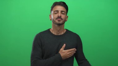 Direk bakışları olan İspanyol genç adam elini göğsüne koyar ve yeşil bir stüdyoda ileriye bakar; samimiyet.