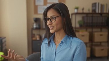 Mavi gömlekli kadın, arka planda raflar ve bitkilerle bir ofis ortamında konuşurken gülümsüyor ve profesyonel bir iç işyeri atmosferi yaratıyor..