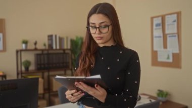 Modern bir ofiste doküman okuyan bir kadın, duvarında çizelgeler ve raflarda bitkiler olan, profesyonel bir işyeri dekorasyonu sergiliyor..