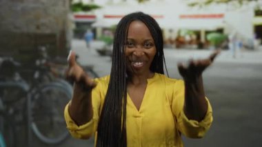 Kentsel sokak ortamında gülümseyen sarı tişörtlü, arka planda bisikletli bir kadın şehir ortamında güneşli gökyüzünün altında neşe ve mutluluk taşıyor..