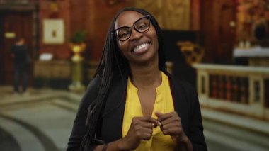 Gözlüklü Afrikalı Amerikalı bir kadın arka planda zarif bir dekor olan bir kilise ortamında neşeyle gülümsüyor..