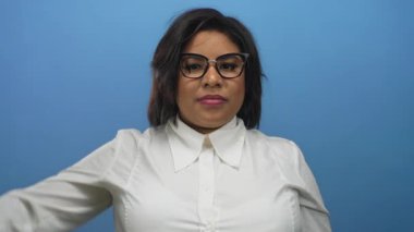 Kendinden emin bir ifadeyle aşağıya bakan, mavi arka planda gözlüklü ve beyaz gömlekli bir kadın, güçlendirilmiş bir Latin kimliği tasvir ediyor..