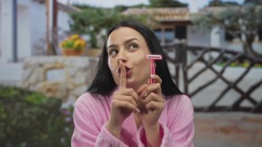 Pembe bornozlu kadın elinde jiletle evinin önünde neşeli bir ifadeyle güneşli bir günde kırsal dekor ve yemyeşil bir şeyle şşş yapıyor..