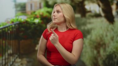 Kırmızı gömlekli kadın şehir sokağında dikiliyor. Düşünceli ve gösterişli görünüyor. Arka planda canlı yeşillik ve şehir mimarisi var..