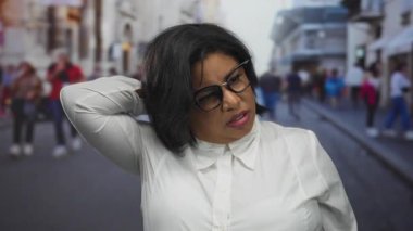 Şehir caddesinde beyaz gömlek giyen Latin bir kadın, boyun ağrısı var gibi görünüyor, kentsel arka plan birden fazla yayanın hareket ettiğini gösteriyor..