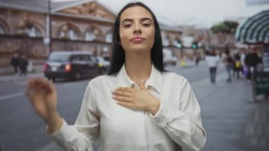 Yoğun şehir ortamında beyaz gömlekli, samimi ve kendine güvenen bir kadın, arka planda insanlar ve arabalarla dolu bir sokakta yemin jesti yapıyor..