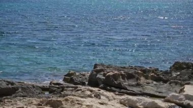 Akdeniz, güneşli gökyüzünün altında, kayalık plaj önplanında, sakin ve pitoresk bir açık hava ortamının özünü yakalıyor..