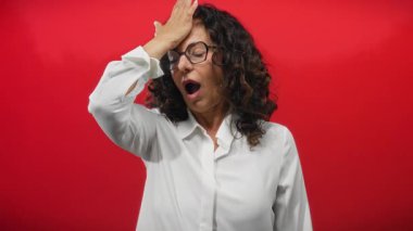 Kıvırcık saçlı bir kadın canlı kırmızı bir arkaplan karşısında şaşkınlığını dile getiriyor, el alında, gözlük takıyor ve beyaz gömlek giyiyor, duygularını gösteriyor ve olgun cazibesini sergiliyor..