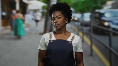 Önlük giymiş bir kadın şehir caddesinde arka planda bulanık duruyor. Değişik şehir yaşamlarını ve hareketli trafiği gösteriyor..