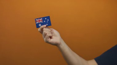 Turuncu arka planda Avustralya bayrağı taşıyan bir adam. Milliyeti ve kimliği vurgulayan minimalist bir yapıda, canlı bir duvarın üzerinde izole edilmiş bir el..