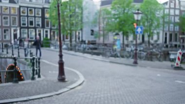 Amsterdam 'daki Bulanık Sokak Sahnesi. Kanal kenarındaki kaldırım taşlı bir yolda Bokeh dolu arka planda dağılmış yayalar ve bisikletçiler var..