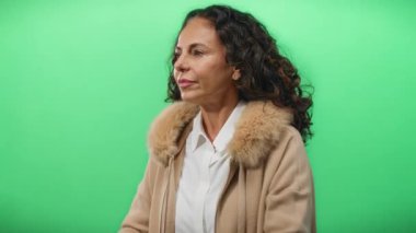 Kıvırcık saçlı bir kadın yeşil arka plan karşısında dürbün hareketi yapıyor, bej bir ceket giyiyor, oyuncu ve yaratıcı bir ortam öneriyor..