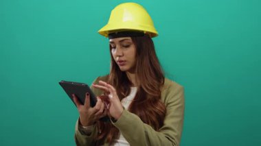 İspanyol kadın mimar yeşil arka plan duvarına başlık takarken tablet kullanıyor, mühendislik görevlerine odaklanıyor, mesleki becerilerini ve özverisini sergiliyor.
