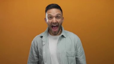 Canlı turuncu arka plana karşı sevinç ifade eden genç adam, geniş bir gülümseme içinde günlük gri gömlek giyerken, dinleyicileri enerjik ve coşkulu jestlerle cezbediyor.