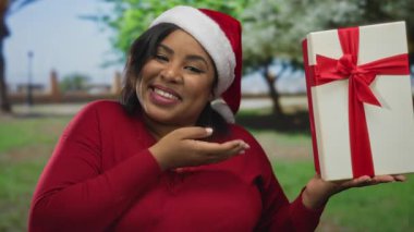 Noel Baba şapkalı kadın, elinde hediyeyle parkta gülümsüyor. Mutluluk ve tatil ruhunu gözler önüne seriyor.