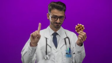 Steteskoplu genç İspanyol doktor mor stüdyoyu işaret eden waffle ve parmak tutuyor; sağlıklı mizah.