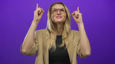 Kadın gülümsüyor ve iki eliyle yukarı doğru işaret ediyor canlı mor bir arka plan, gözlük ve bej bir ceket giyiyor, enerjik ve neşeli bir an yakalıyor.