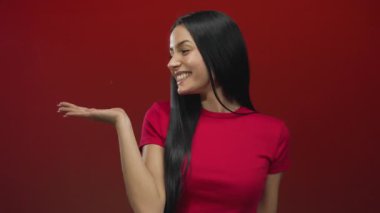 Genç İspanyol kadın, kırmızı izole bir arka plan üzerinde olumlu bir jest ile gülümsüyor, neşeli bir ifadeyle kendine güveni ve pozitifliği yayıyor ve şık siyah saçlı, canlı giyinmiş..