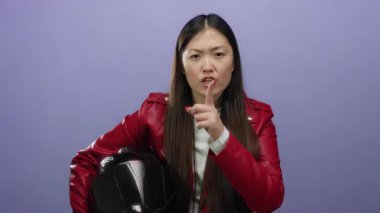 Kırmızı ceketli kadın miğfer tutarken mor arka planda sessizliği öneriyor. Dudaklarında parmaklarıyla poz veren genç Asyalı bir kadın var..